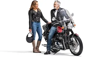 Sitting on a parked black bike, a grey-haired man turns to speak to a woman in a black leather jacket. She holds her helmet.